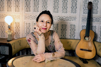 Eine Frau sitzt an einem Tisch. Sie stützt den Kopf mit ihrer Hand ab und lächelt leicht. Im Hintergrund steht links neben ihr eine Lampe und rechts von ihr lehnt eine Gitarre an der Wand, welche ebenfalls mit auf der Sitzbank vor dem Tisch steht.