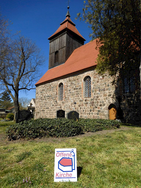 Kirche aus grauen Steinen, einem dunkel braunen Turm und roten Dächern. Vorne steht ein weißes Schild, das mit roter und blauer Schrift sagt "Offene Kirche"