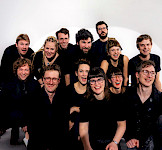 Eine Gruppe aus 13 schwarz gekleideten, jungen lachenden Menschen posieren vor einer weißen Wande